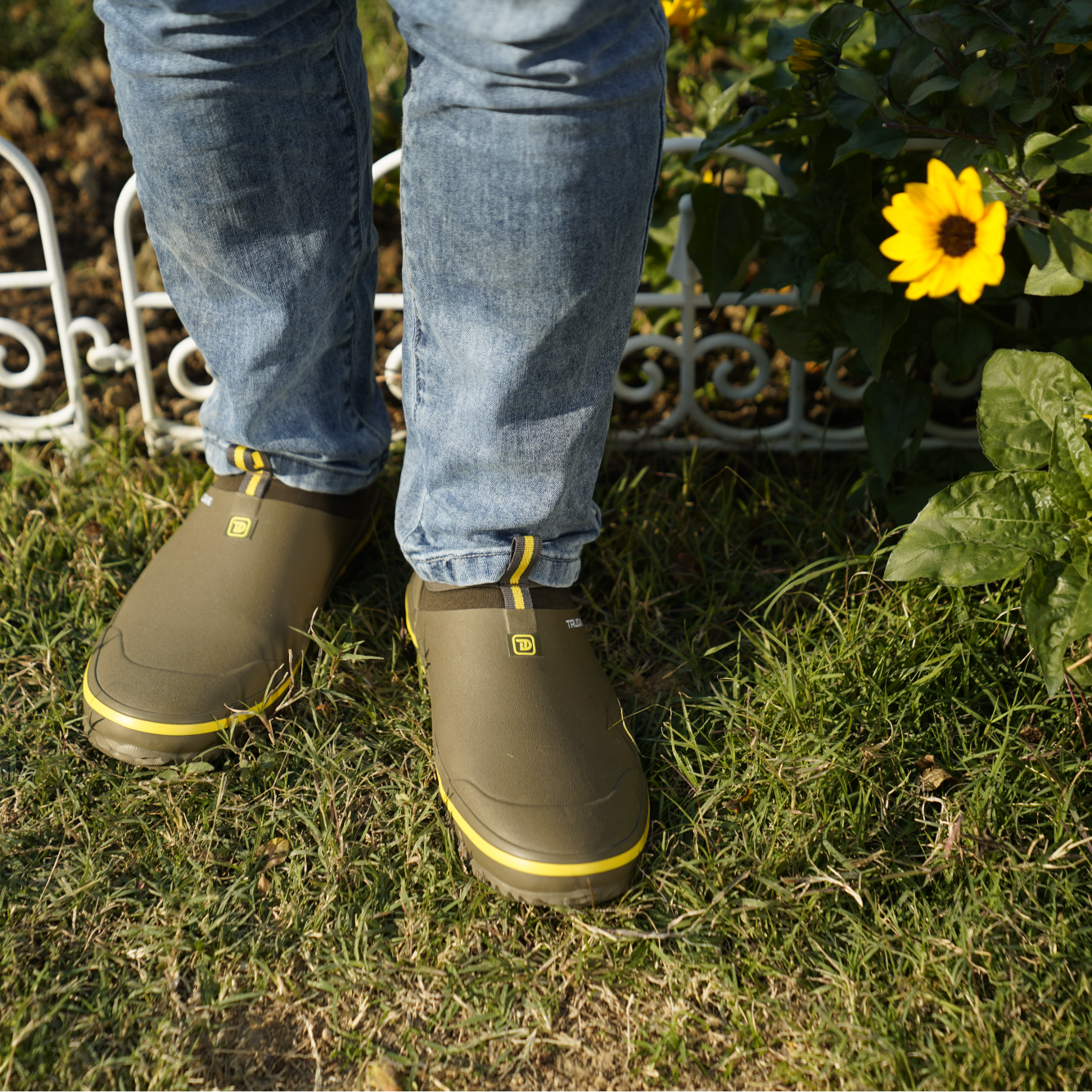 alt="Lifestyle image of Trudave GreenStep Series waterproof slip-on garden shoes in olive green color with yellow trim, worn outdoors while gardening near flowers, showing durable rubber design, comfortable fit, and waterproof protection for yard and garden work"